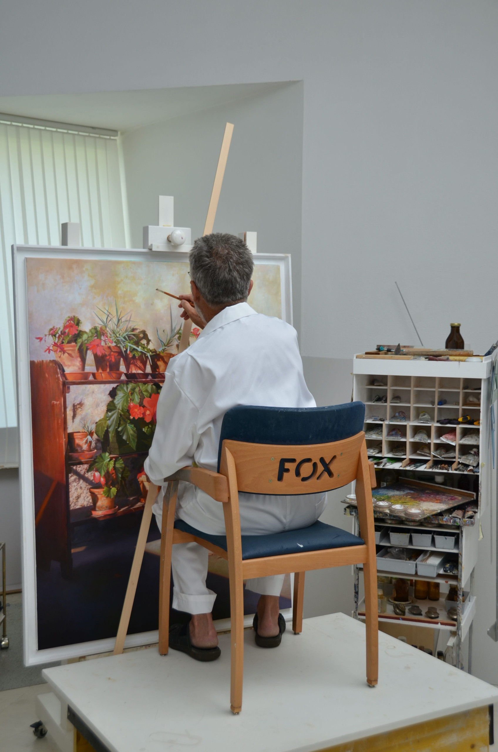 Pavel Florjančič painting a still life of potted flowers on an easel in his art studio, wearing a white painter’s coat.